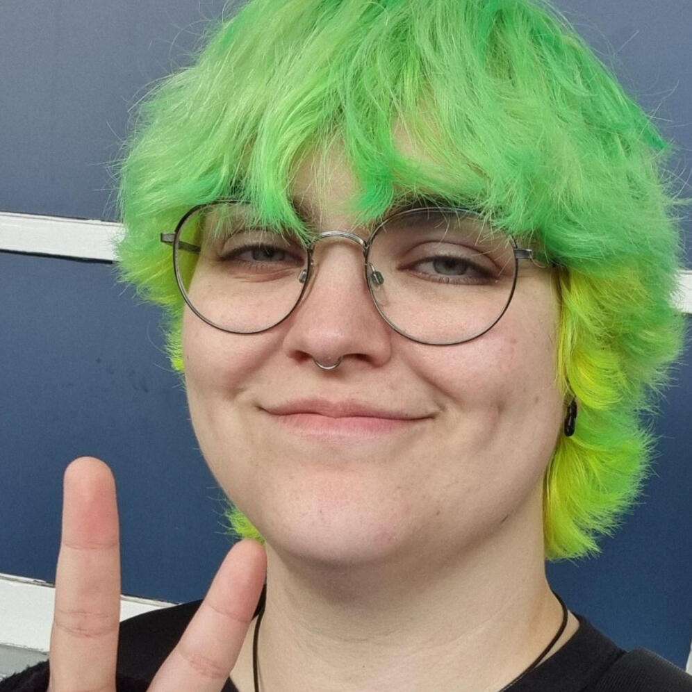 A picture of Ellis smiling at the camera. They are light skinned with short, brown curly hair and grey-green eyes. They're wearing round silver glasses, a green knitted scarf, and a pair of headphones, and they have septum and eyebrow piercings.